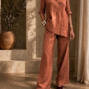 Op de afbeelding: Een roestkleurige linnen set bestaande uit een tuniek met V-hals en ellebooglange mouwen en een bijpassende broek met wijde pijpen. De tuniek heeft een asymmetrische zoom en de broek heeft een relaxte pasvorm. De stof heeft een zichtbare textuur.
