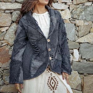 May include: A dark gray jacket featuring a patchwork design with varied textures, frayed edges, and three light-colored buttons. The jacket is worn over a white top and a cream-colored skirt with a brown geometric pattern printed on the fabric.