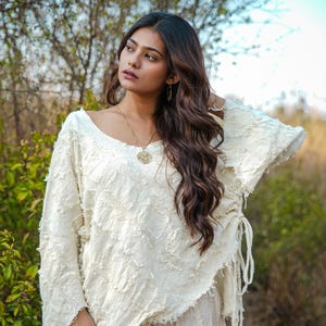 Könnte beinhalten: Ein cremeweißes Langarmshirt mit texturierter, ausgefranster Stoffoberfläche, weitem Ausschnitt, asymmetrischem Saum und seitlichen Kordelzügen. Das Kleidungsstück wird mit einer passenden cremeweißen Hose und einer goldenen Anhängerkette kombiniert.