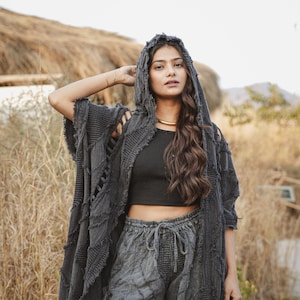 May include: A dark charcoal gray hooded cardigan featuring a textured, patchwork-style fabric with frayed edges and open-weave detailing. The garment is worn over a black sleeveless crop top and matching dark gray drawstring trousers.