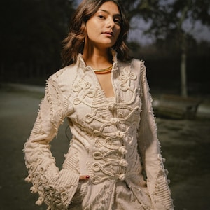 May include: An off-white, long-sleeved jacket featuring intricate rope-style embroidery and decorative knot closures down the front. The garment has a high collar, frayed edges, and textured fabric panels with a structured, fitted silhouette.