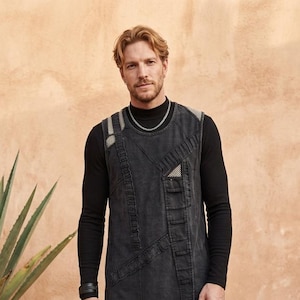 Könnte beinhalten: Eine ärmellose Tunika in Anthrazit mit hohem Ausschnitt, mehreren vertikalen Steppnähten, Netztaschendetails und einem langen Saum. Das Kleidungsstück wird über einem schwarzen Langarmshirt und einer schwarzen Cargohose mit Lederstiefeln getragen.