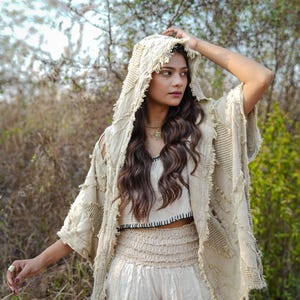 May include: A beige, three-piece outfit featuring a hooded jacket with fringed edges, a crop top, and high-waisted pants. The jacket has open sleeves and a textured design. The crop top has a black trim, and the pants have a gathered waist. The outfit is made of a light, natural fabric.