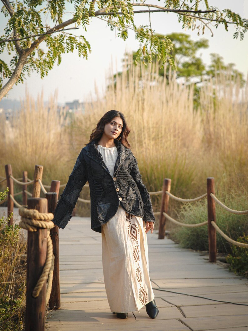 May include: A dark charcoal gray textured jacket with a notched collar and three light-colored buttons, worn over a cream-colored long dress featuring a vertical brown geometric pattern along the front panel and black pointed-toe ankle boots.
