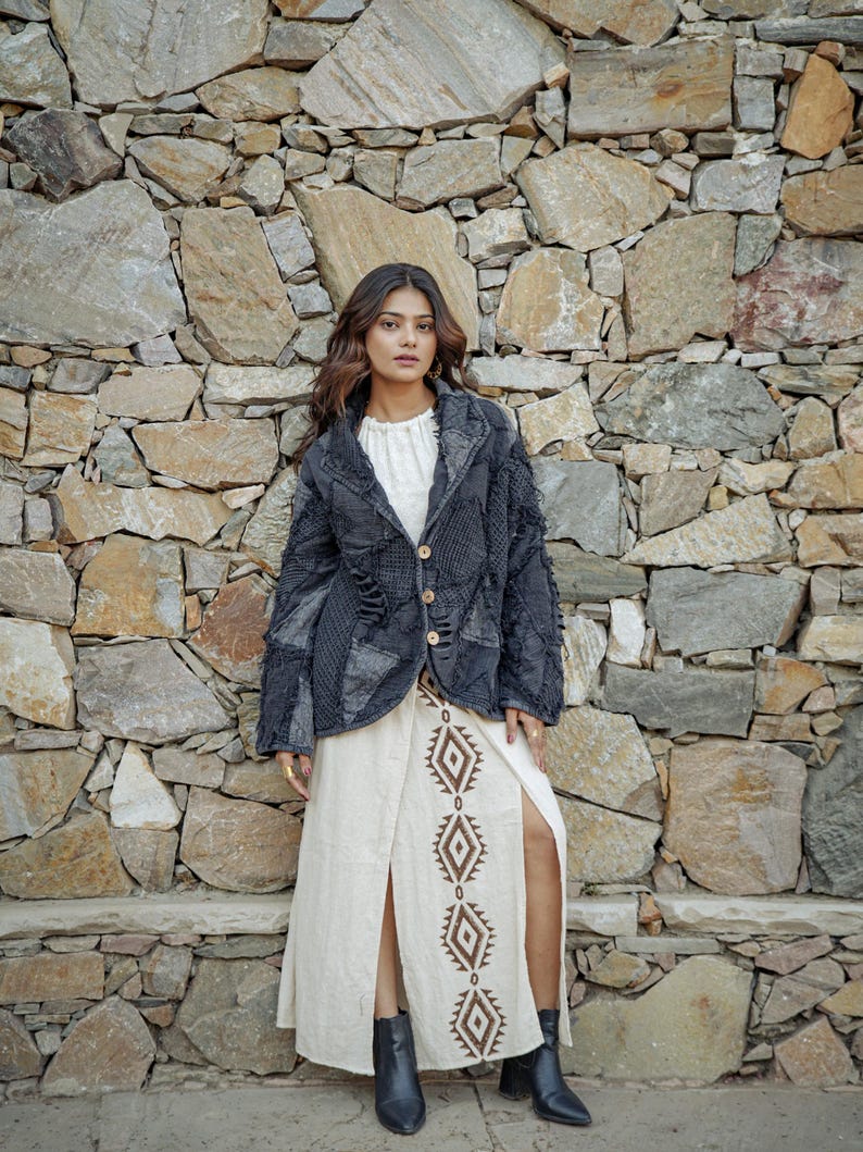 May include: A charcoal gray patchwork blazer with frayed edges and three light-colored buttons, worn over a cream-colored long skirt featuring a vertical brown diamond pattern and a high front slit, paired with black ankle boots.