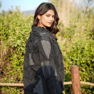 May include: A dark charcoal gray jacket featuring a patchwork design with various fabric textures, frayed edges, and visible stitching. The garment has a collared neckline, long sleeves, and a button-down front, worn over a light-colored bottom garment.