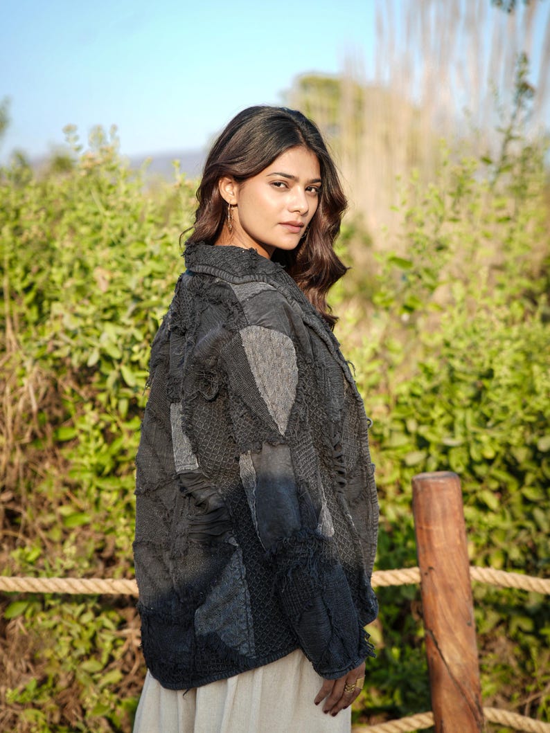 May include: A dark charcoal gray jacket featuring a patchwork design with various fabric textures, frayed edges, and visible stitching. The garment has a collared neckline, long sleeves, and a button-down front, worn over a light-colored bottom garment.