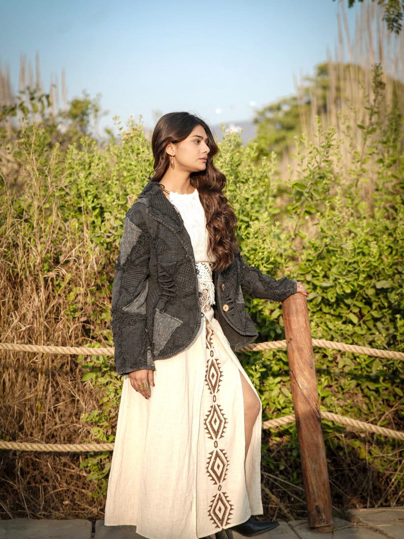 May include: A dark charcoal gray patchwork jacket with distressed textures and visible stitching worn over a cream-colored maxi skirt featuring a vertical row of brown diamond-shaped geometric patterns and a high side slit along the leg.