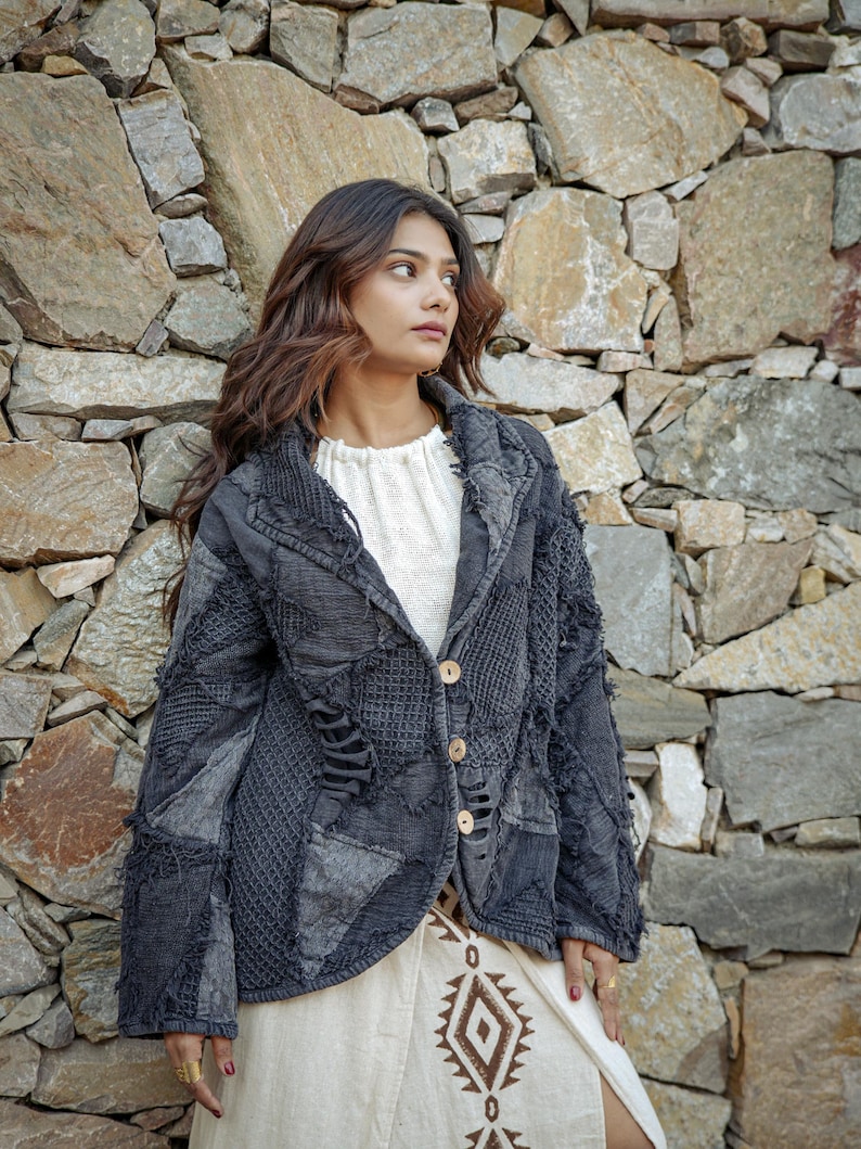 May include: A dark gray jacket featuring a patchwork design with varied textures, frayed edges, and three light-colored buttons. The jacket is worn over a white top and a cream-colored skirt with a brown geometric pattern printed on the fabric.