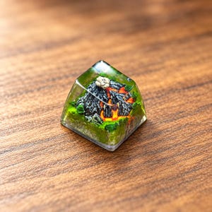 May include: A square mechanical keyboard keycap featuring a miniature 3D volcanic landscape encased in clear resin. The design displays a dark grey mountain with bright orange lava flows, a small white smoke plume, and green foliage.