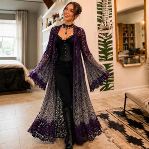 May include: A long, open-front crochet duster cardigan featuring a gradient colour transition from deep purple at the shoulders and hem to a light grey in the centre. The garment has wide, flared sleeves and a detailed, intricate lace-like pattern.