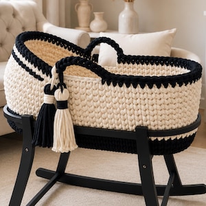 May include: A cream-colored crocheted baby bassinet with a thick black crocheted rim and handles. The bassinet features two large decorative tassels in black and cream hanging from the side and rests on a black wooden rocking stand base.