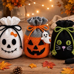 May include: Three crocheted drawstring bags featuring Halloween themes: a white skull with a black bow, an orange pumpkin with a gray top and a small white ghost charm, and a black cat with green eyes, a pink nose, and a long tail.