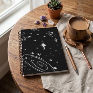May include: A black spiral-bound notebook featuring a white hand-drawn illustration of a galaxy, various stars, and celestial shapes. The notebook is positioned on a wooden surface alongside a wooden pen, a wooden mug, and a small succulent plant.