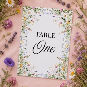 May include: A rectangular white table sign featuring the black text TABLE One in the centre, surrounded by a decorative border of colourful wildflowers including daisies, lavender, and clover, printed on a smooth cardstock material.