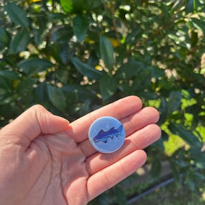 Op de afbeelding: Een ronde, lichtblauwe button met een haaienillustratie. De haai is donkerblauw met witte accenten en de achtergrond heeft kleine witte bellen. De button wordt in een hand gehouden, tegen een achtergrond van groen gebladerte.