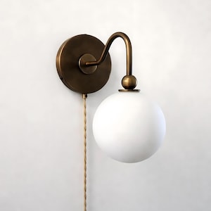 May include: A wall-mounted light fixture featuring a circular antique brass backplate and a curved arm that supports a spherical, frosted white glass globe shade. A beige twisted fabric cord hangs vertically from the base of the fixture.