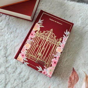 Op de afbeelding: Een hardcover boek getiteld Rebecca van Daphne du Maurier met een dieprode kaft, een gouden illustratie van een sierlijk hek en roze bloemaccenten. Het boek ligt naast een oranje boek en een donkerrood boek met een zwarte boekenstandaard.