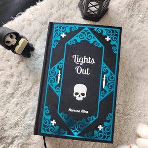 Può includere: Un libro con copertina rigida nera intitolato Lights Out di Navessa Allen, caratterizzato da un bordo ornato blu brillante, icone di pugnali bianchi e un teschio bianco centrale. Una piccola statuetta del tristo mietitore e una lanterna in metallo nero sono vicine.
