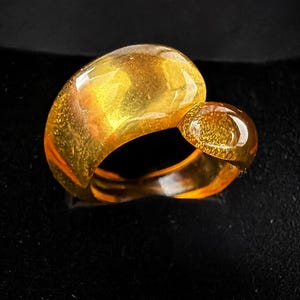 May include: A translucent amber ring featuring a smooth, polished surface with a warm golden-orange hue. The design consists of a wide, rounded band with a smaller, distinct circular accent piece attached to the side, creating a continuous, organic shape.