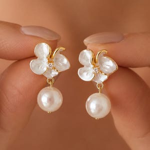 May include: A pair of gold-tone earrings featuring white, petal-shaped floral elements with a small clear stone at the center. Each earring is finished with a dangling, round, white pearl-like bead suspended from the base of the flower design.