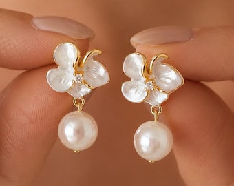 Può includere: Un paio di orecchini color oro con elementi floreali bianchi a forma di petalo e una piccola pietra trasparente al centro. Ogni orecchino è completato da una perla bianca rotonda pendente fissata alla base del motivo floreale.