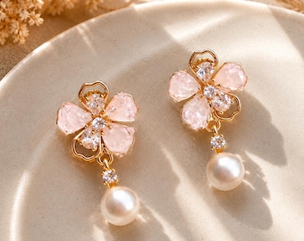 May include: A pair of gold-tone earrings shaped like flowers with light pink translucent petals, clear sparkling stones at the center, and a single white pearl dangling from the bottom of each flower, secured by a small clear stone connector.