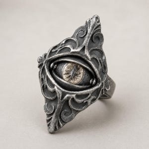 May include: A silver-toned metal ring featuring an elongated, pointed shape with intricate scrollwork patterns. The center of the ring displays a detailed, realistic eye motif with a dark iris and a light-colored, textured pupil set within the metal.