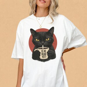 May include: White t-shirt featuring a black cat illustration holding a coffee mug with the text "IS HE DEAD YET?" The cat is set against a red circle. The shirt has short sleeves and a relaxed fit. The StreetInk Tees logo is visible.