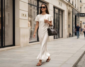Pode incluir: Um vestido longo de cetim na cor creme com mangas curtas esvoaçantes e decote redondo. O visual inclui uma bolsa de mão de couro trançado em marrom escuro, sandálias rasteiras de couro marrom e óculos de sol pretos.