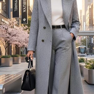 Könnte beinhalten: Ein langer, zweireihiger Mantel aus einer grauen Wollmischung über einem weißen Rollkragenpullover und einer passenden grauen Hose mit weitem Bein. Ein schwarzer Ledergürtel mit goldener Schnalle, schwarze Lederstiefeletten und eine strukturierte schwarze Handtasche vervollständigen das Outfit.