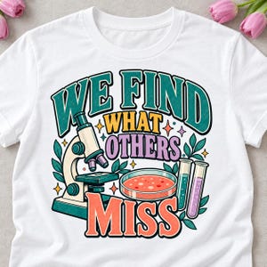 May include: A white t-shirt featuring a colourful graphic design with a microscope, a petri dish, and two test tubes. The text reads "WE FIND WHAT OTHERS MISS" in bold, multi-coloured lettering, surrounded by small star and leaf illustrations.