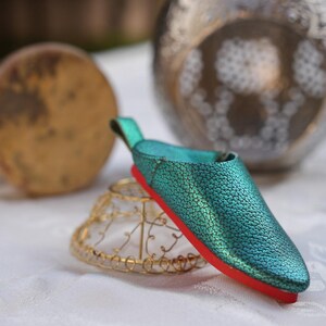 Könnte beinhalten: Ein türkisfarbener Lederschuh mit roter Sohle ruht auf einem goldenen Drahtständer. Der Schuh hat eine strukturierte Oberfläche und eine kleine Schlaufe an der Ferse. Weitere dekorative Gegenstände befinden sich im Hintergrund.