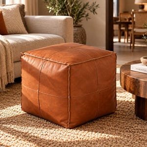 May include: A square, cognac-coloured leather ottoman featuring a smooth, matte finish and prominent stitched seams that create a grid pattern across the top and sides. The piece has a structured, boxy shape with visible piping along the edges.