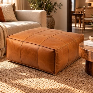 May include: A square, low-profile ottoman upholstered in tan leather with a grid-like pattern of stitched seams across the top and sides. The piece features a smooth, matte finish and is constructed with structured, upright edges and flat surfaces.