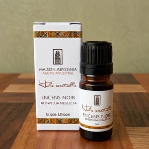Peut inclure: Une bouteille d'huile essentielle et sa boîte sont présentées sur une surface en bois. La bouteille est en verre ambré foncé avec un bouchon noir. La boîte est blanche avec des accents dorés et le texte "ENCENS NOIR BOSWELLIA NEGLECTA" et "Huile essentielle".