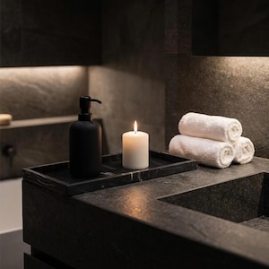 May include: A black soap dispenser and a lit white candle sit on a black marble tray. Rolled white towels are stacked nearby. The items are arranged on a dark gray countertop in a bathroom setting.