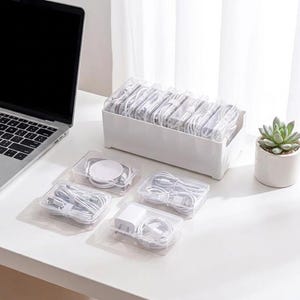 May include: A white rectangular plastic storage box with multiple internal dividers containing coiled white charging cables, alongside four smaller clear plastic cases holding individual white cables and a power adapter.