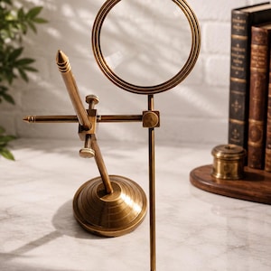 May include: A brass magnifying glass mounted on an adjustable stand with a circular base and a vertical rod. The device features a round lens frame and a pointed metal stylus attached to the central joint. The word LEGENDARY is printed below.