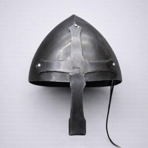 May include: A dark grey steel nasal helmet featuring a pointed crown and a central vertical bar extending down from a horizontal cross-shaped reinforcement piece. The helmet is secured with rivets and includes a thin black leather chin strap.