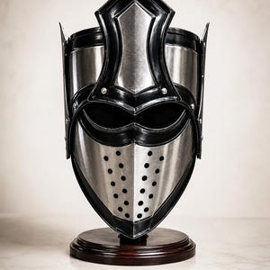 May include: A metallic helmet featuring a polished silver finish with contrasting black trim and a central vertical crest. The faceplate includes two horizontal eye openings and a lower section with a grid of circular ventilation holes.