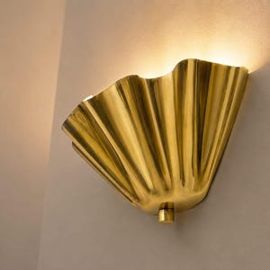 Puede incluir: Un aplique de pared de color dorado con forma de abanico plisado y acampanado. El accesorio de metal tiene una superficie pulida y reflectante que emite un brillo cálido y suave desde la parte superior, proyectando luz hacia la pared.