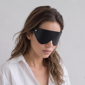 Puede incluir: Un antifaz negro liso con una silueta curva y suave que cubre la parte superior del rostro. La máscara se asegura con un pequeño remache de metal plateado en el lateral, ofreciendo un aspecto limpio que descansa sobre los ojos.