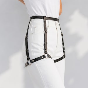May include: A black leather leg harness featuring a waist belt connected to two adjustable thigh straps by vertical leather strips. The harness includes silver-toned metal buckles, rivets, and circular rings, worn over white denim jeans and a white t-shirt.