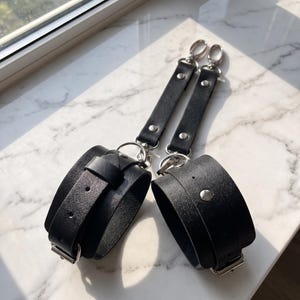 May include: A pair of black leather wrist cuffs featuring adjustable buckle straps, silver-toned metal hardware, and two vertical leather connector straps attached to swivel lobster claw clasps at the top for secure fastening and attachment.