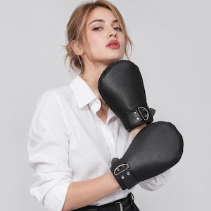 May include: A pair of black leather-textured mittens featuring a wide wrist cuff with a silver-toned metal D-ring and two rivets. The mittens have a rounded, padded shape and a smooth, solid black finish, designed to cover the hands and wrists.