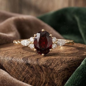 May include: A gold ring featuring a large oval-cut deep red garnet gemstone at the centre, flanked by two smaller round diamonds and two marquise-cut diamonds on each side, set on a twisted gold band with a polished metallic finish.