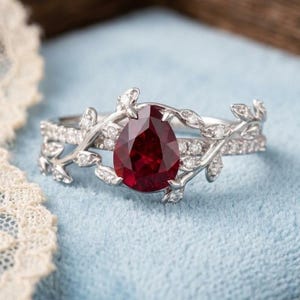 Può includere: Un anello color argento con una pietra preziosa rosso scuro a taglio pera al centro. La fascia è decorata con intricati motivi a foglia in metallo e piccole pietre chiare incastonate a pavé che si estendono lungo i lati, creando un aspetto dettagliato.