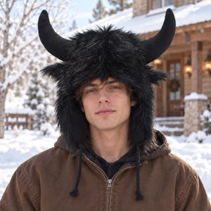 May include: A black faux fur hat featuring two large, curved horns extending from the top. The hat has a shaggy texture, ear flaps that extend down the sides, and two thin braided cords hanging from the bottom of the ear flaps for fastening.