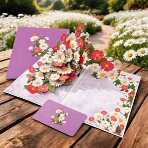 Peut inclure: Une carte de vœux pop-up présentant un bouquet en relief de marguerites blanches et de fleurs rouges, accompagnée d'une enveloppe violette et d'une petite carte rectangulaire, ornées d'illustrations florales sur un fond violet clair.